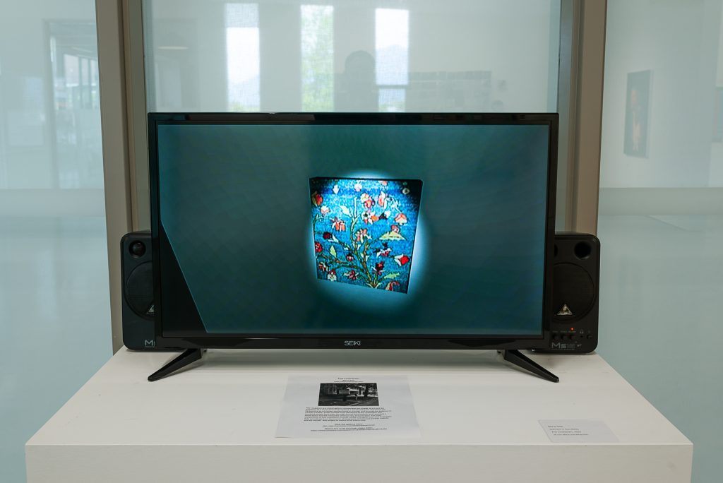 Installation of a TV and two speakers mounted on a plinth. On the TV is an image of a stained-glass tile of flowers with a spotlight shining on it. 