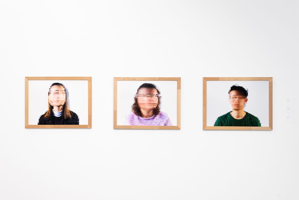 Three wall-mounted prints in light brown frames of portraits with pixelated, blurry faces. The left subject has long hair and a black shirt; the middle subject has brown hair and purple glasses and shirt; and the right subject has short black hair and an emerald green shirt. 