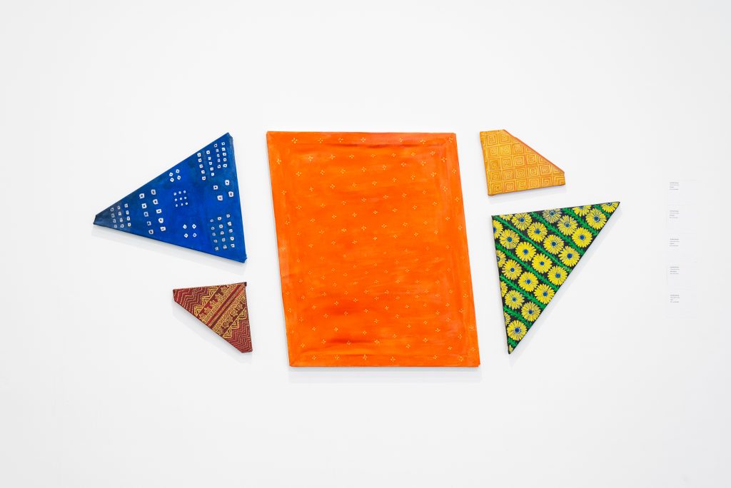 A group of triangles and polygons mounted onto the wall, wrapped tightly with brightly coloured, patterned fabric in shades of orange, blue, red and yellow. 