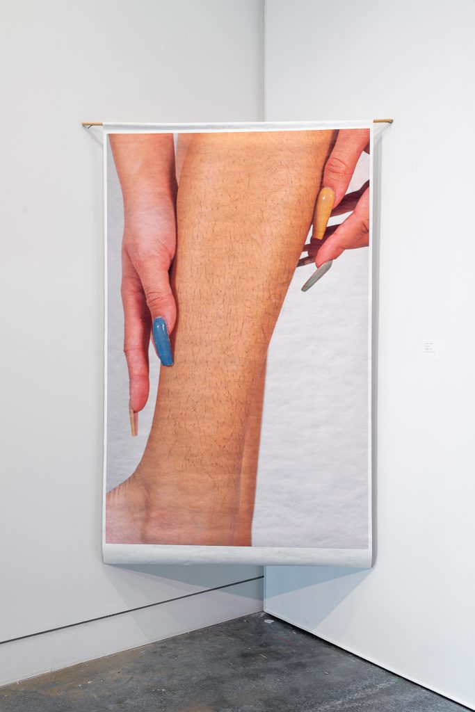 Dowel-mounted print of a leg calf with stubble, being stroked with hands that have long painted fingernails. 