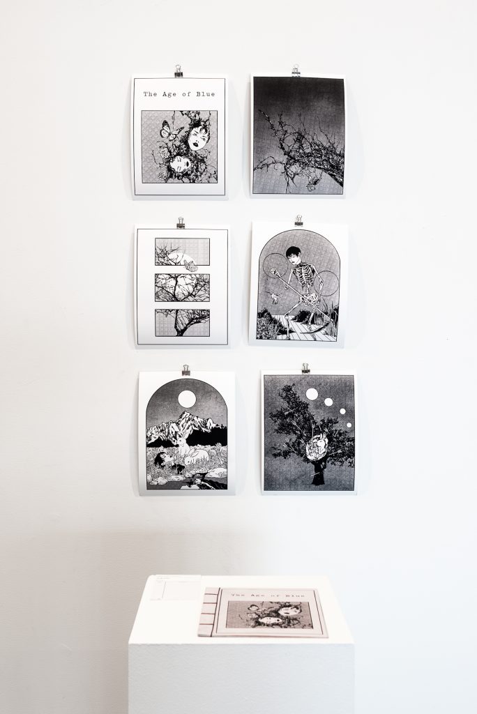 A zine, “The Age of Blue”, sits on a plinth with a cover drawing of of two heads emerging from a tangle with butterflies emerging; on the wall behind hang six prints including the cover image, featuring images of trees, human figures and animals. 
