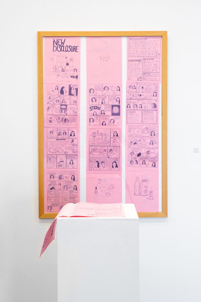 A pink zine with purple ink titled “New Disclosure” lays open on a plinth; on the wall behind, in one large light brown frame are the pages of the zine laid out. 