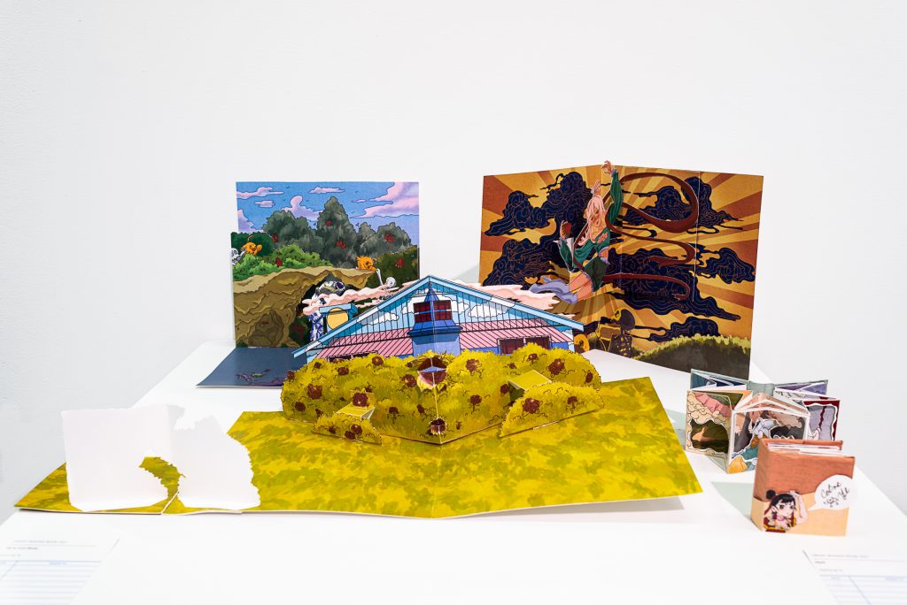 Three pop-up cards and two small books arranged on a plinth, depicting illustrations of a house peeking behind bushes, colourful tree-lined landscapes, and human figures. 