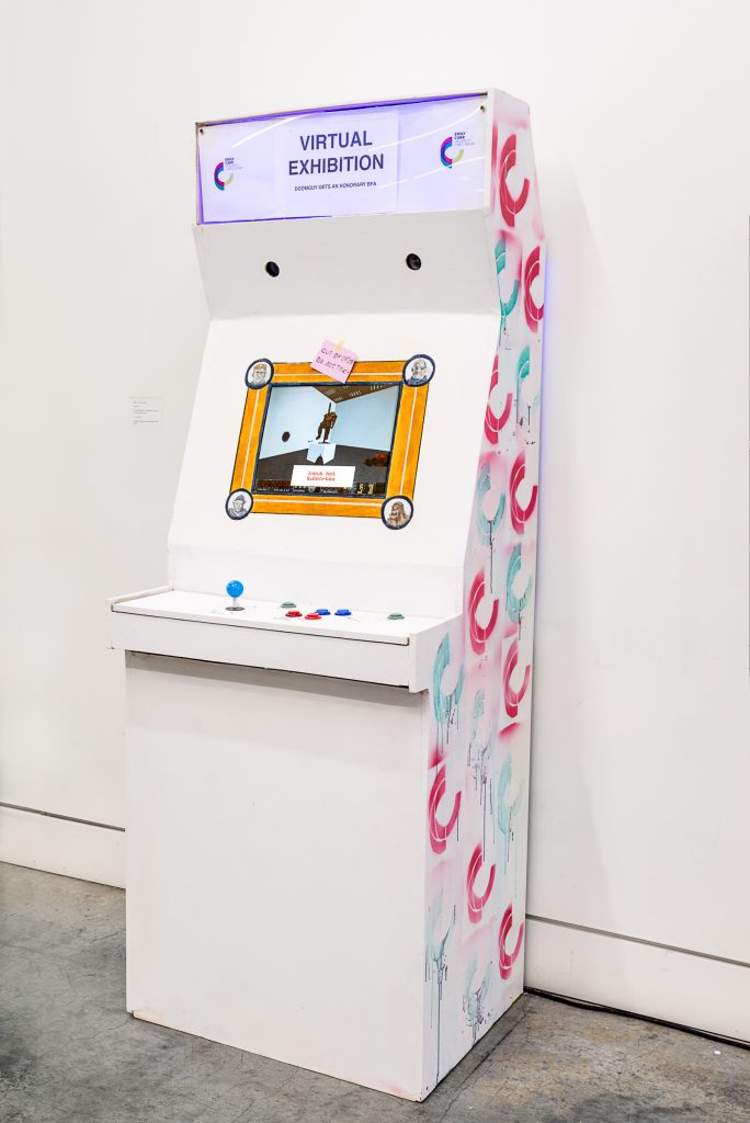 A white arcade game console with the title “VIRTUAL EXHIBITION: DOOMGUY GETS AN HONORARY BFA” with the Emily Carr University logo spray-painted in pink and turquoise on the side. The console’s screen shows an exhibition space with a message saying “Input Not Supported,” and a pink sticky note is tacked on the console reading “Out of Order / Do Not Touch.” 
