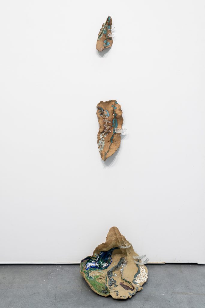 Brown clay sculptures arranged vertically, with two mounted on the wall and one on the ground. They all have a thin, rippled shape like a wrinkled leaf, and shards of textured glass and splatters of crackled white, blue and green paint on them. 
