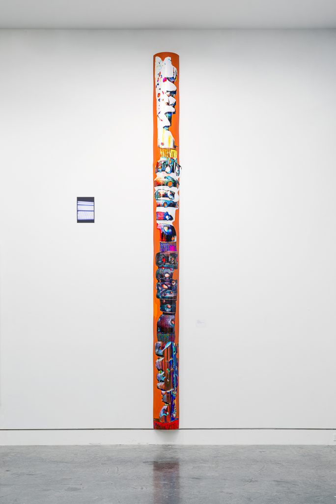 Brightly coloured, abstract corrupted images are cut out and affixed along the length of on a tall, thin orange plank mounted to a wall. 