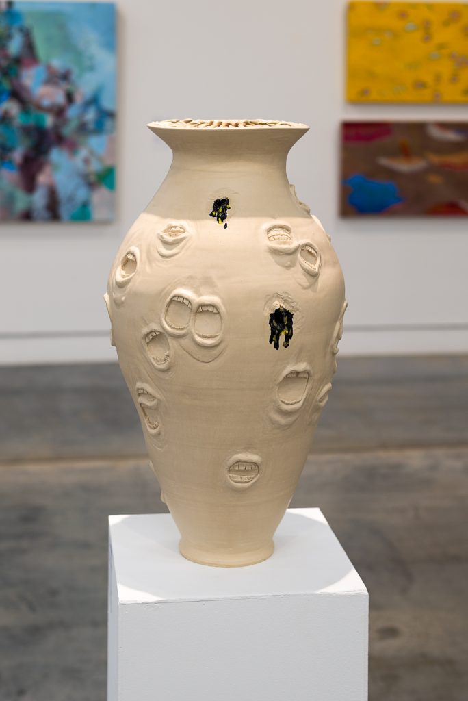 Ceramic vase on a plinth with a narrow base, wide middle and narrow spout with a lip. The surface is dotted with many open mouths in relief, and some open orifices with black paint drip out. 