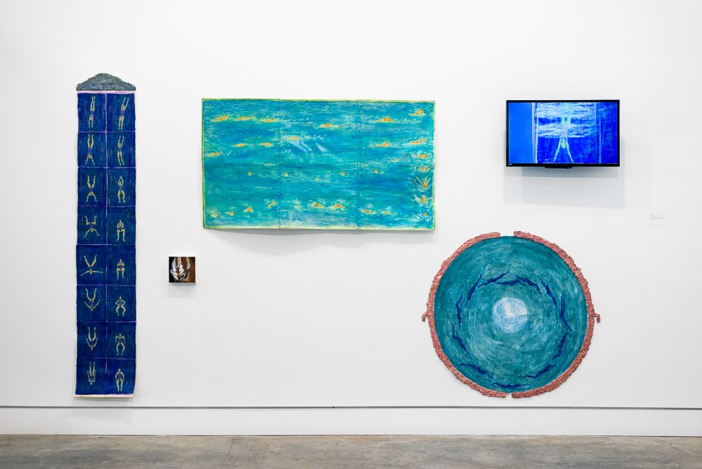 Installation of wall-mounted objects: A long length of dark blue fabric with small green human figures; a small painted tile; a wide, textured teal painting, a circular teal painting with dark blue strokes and brown border; and a small TV showing a bright blue image of a figure similar to the pattern on the fabric. 