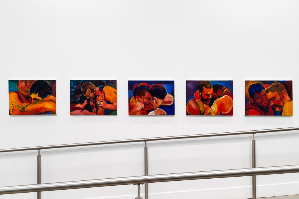 Five portraits mounted above metal railings. Each depicts the tightly cropped heads of two men wrestling, rendered in bright shades of orange and red with blue backgrounds. 
