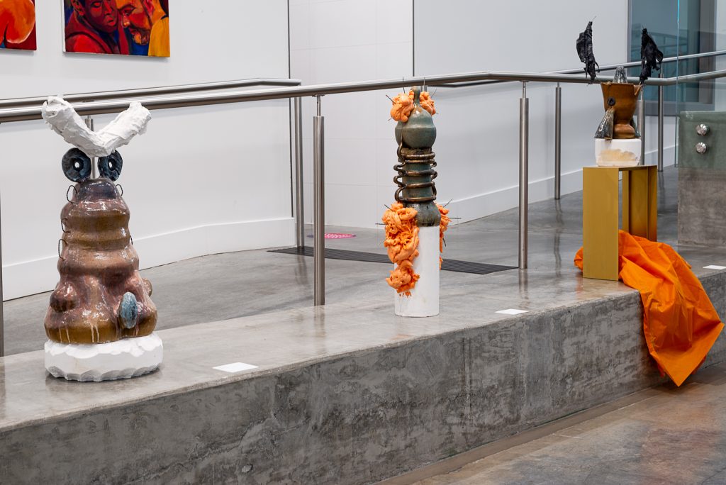 Three sculptures of ceramic vessels. The left is brown and has white plaster antennae; the middle is grey and has gloopy orange foam and nails emerging from it; the right is brown and white, mounted on a wooden box with dripping black forms suspended atop it and an orange plastic tarp below. 