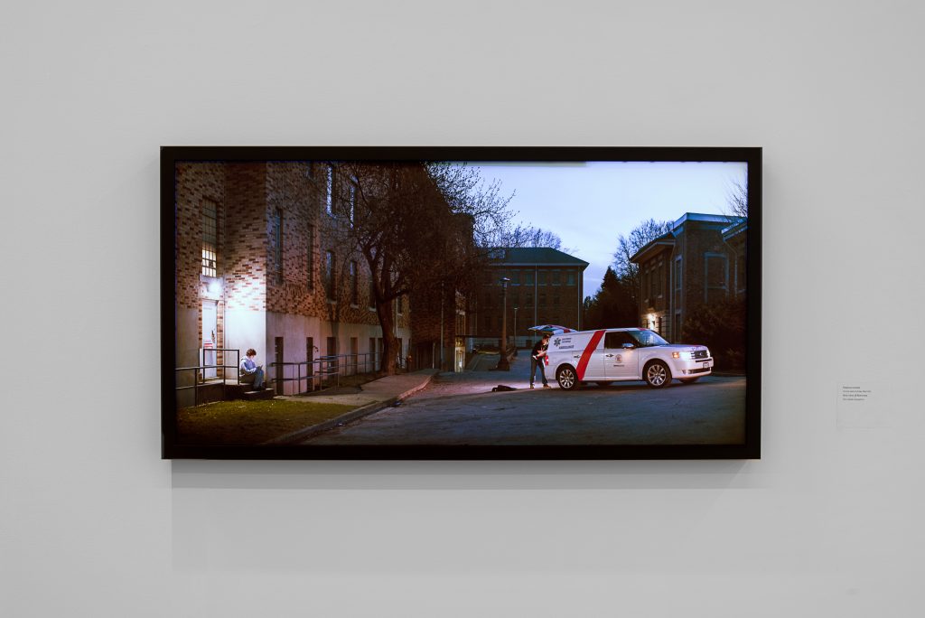 Black-framed photograph of a residential block at night. A person stands in front of the open trunk of an ambulance van, and feet are sticking out from the trunk. To the left is a person sitting on a lit stoop of an apartment building, looking at their phone. 