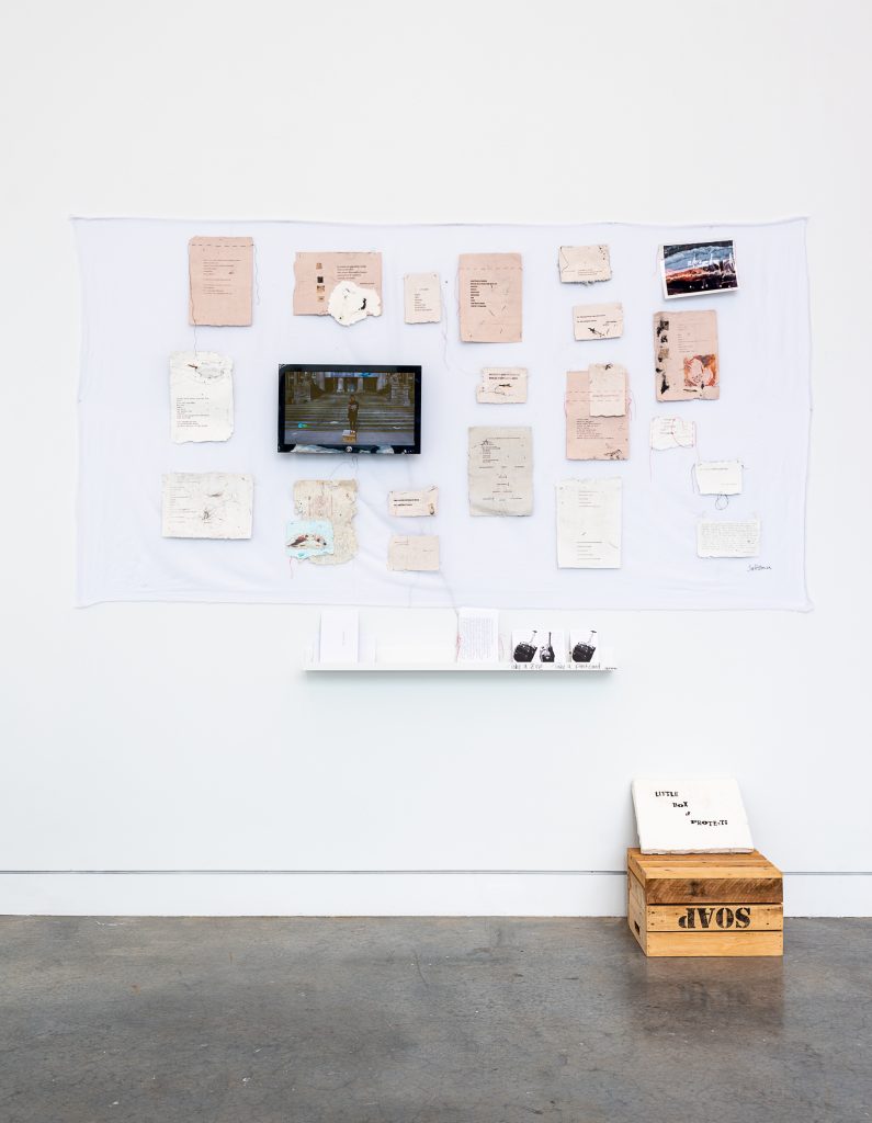 Wall-mounted installation of many pieces of fibrous paper and pictures tacked up against white fabric. The papers have type-written notes and ink drawings on them. On the ground sits an upturned wooden soapbox, and a placard that reads “Little box of protests.” 