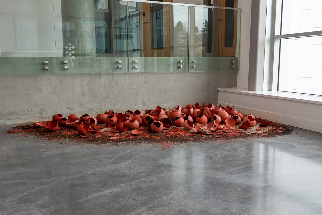 Installation in the corner of a room of a pile of many large, cracked red eggshells. 