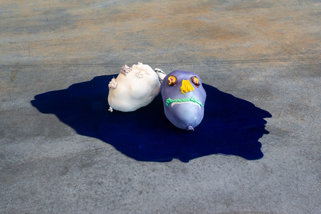 Floor installation of two small head figures made of socks, with squirts of oil paint used to create facial features. One is white and the other is an iridescent blue-grey with yellow nose and green mouth. They rest on top a reflective blue-black surface shaped like a puddle. 