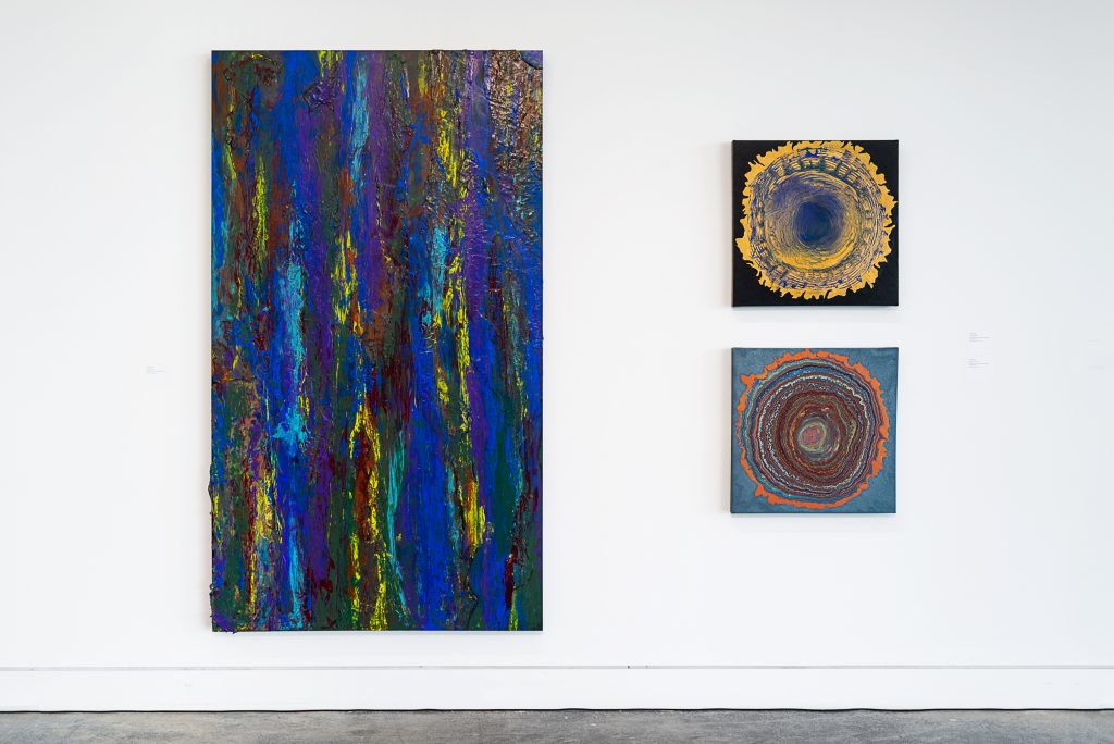 Three abstract paintings; one is large and has vertical brushstrokes of cobalt blue, yellow, and dark green. The others are smaller, square and show tree ring-like formations; the top one has yellow and blue paint on a black background and the bottom one has brown and orange paint on a teal background. 