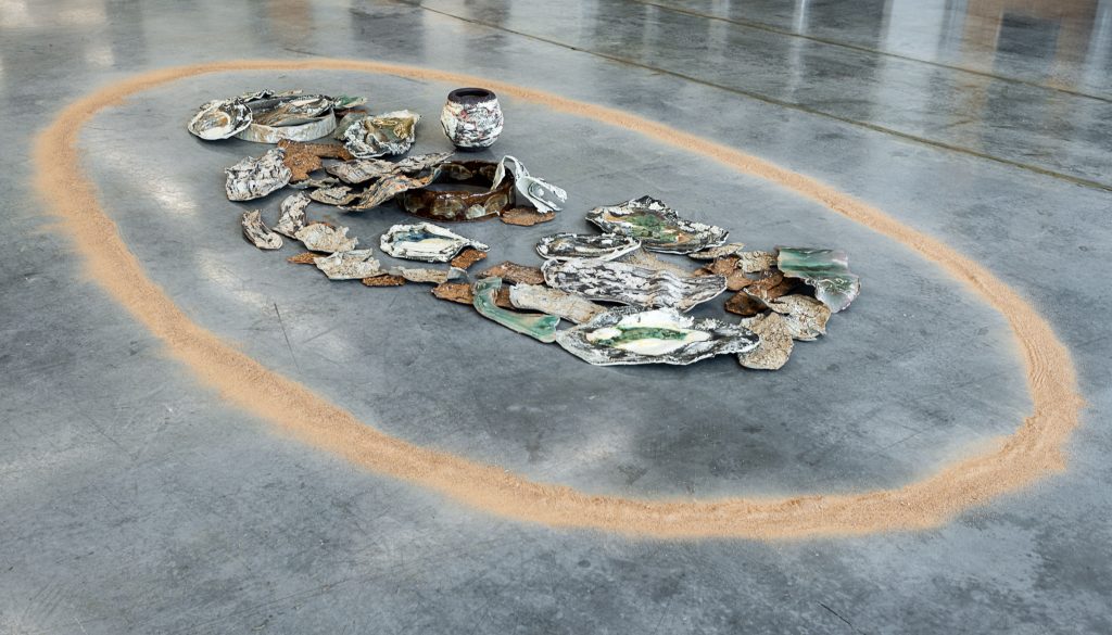 Floor installation of irregularly shaped, flat ceramic pieces covered in paint-strokes of green, blue and brown, arranged in a somewhat overlapping pile with a matching pot beside them. Surrounding the pile is a large ring of orange-brown powder. 