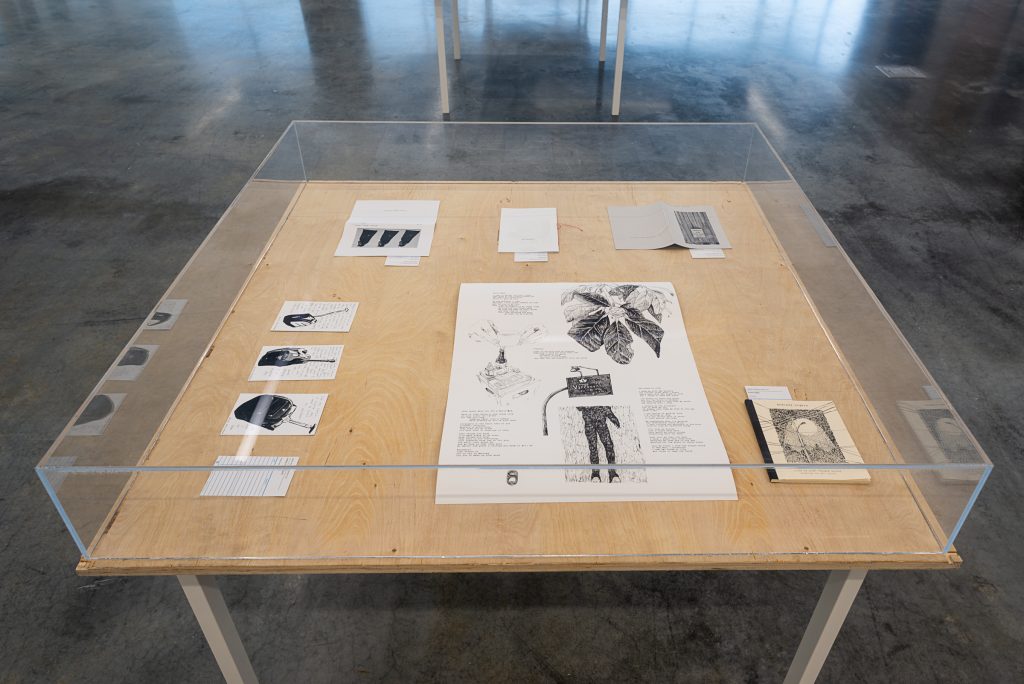 A series of prints and small zines printed on gray and cream paper arranged in a wooden, glass-topped display box. 