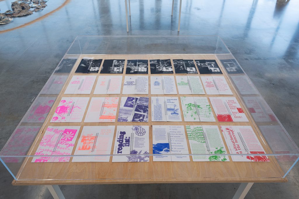 A series of zines, each using a different bright colour of ink on white paper, arranged in a grid on a wooden, glass-topped display box. 
