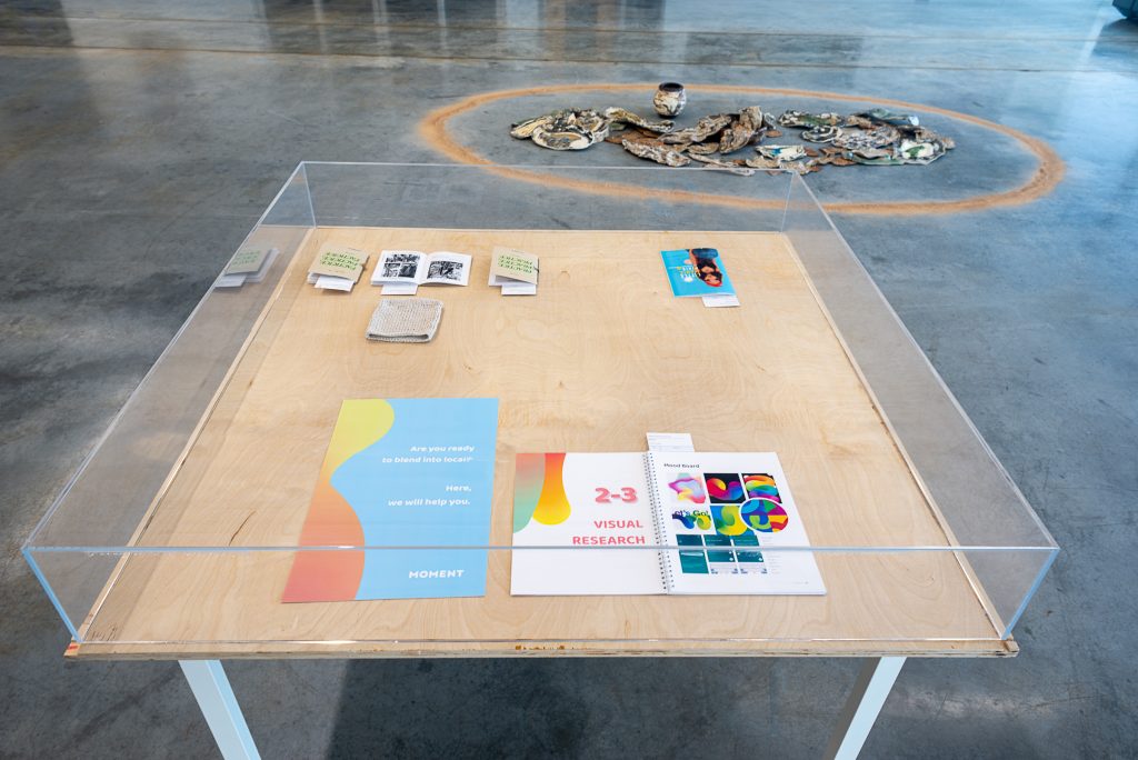 Several groups of small posters and booklets arranged in a grid on a wooden, glass-topped display box. In the background is an installation on the floor of many flat, textured ceramic pieces encircled by a ring of brown-orange powder.