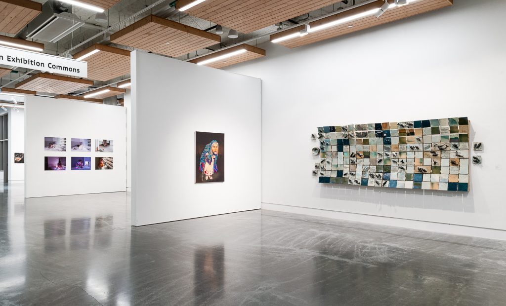 Exhibition installation inside the Emily Carr University building. In the foreground is a mosaic of many square tiles in shades of blue, teal, and brown, and on the adjacent wall to the left is a painting of a figure hunched over wearing a mask and a plaid blanket over their head. On the far wall in a separate area is a series of six purple-toned photographs. 