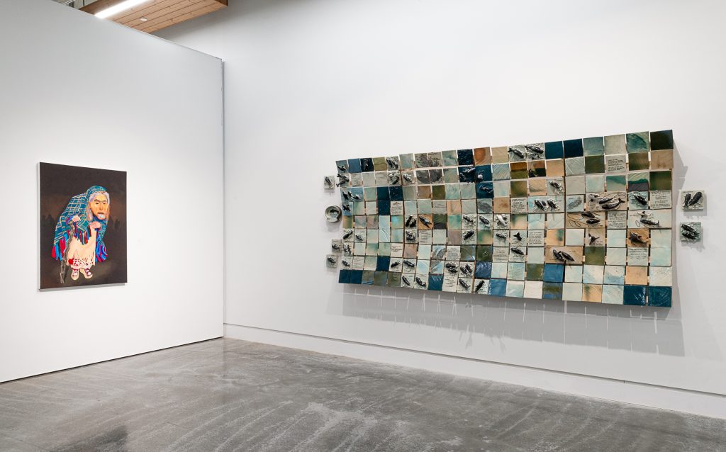 Exhibition installation; in the foreground is a mosaic of many square tiles in shades of blue, teal, and brown, and 3-dimensional whale figures stick out from the surface of several tiles. On the adjacent wall to the left is a painting of a figure hunched over a walking stick, wearing a mask with an iridescent labret inlay and a plaid blanket over their head. 