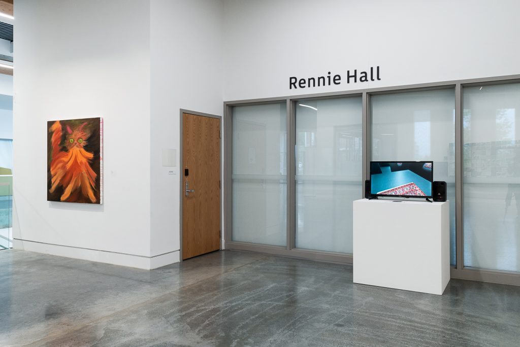 Outside of a room labelled “Rennie Hall,” a TV is mounted on a plinth. On the screen is a teal-coloured floor with the corner of a woven rug poking out. To the left on a wall is a painting of an orange cat with large green eyes and pink ears, painted in large abstract brushstrokes. 
