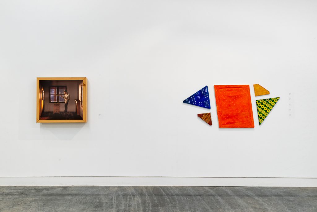 On the left of a wall is a wood-framed photograph of a person in a dimly lit room trimming a bonsai tree. To the right is a group of triangles and polygons mounted on the wall, wrapped with patterned orange, blue, red, yellow and green fabric. 