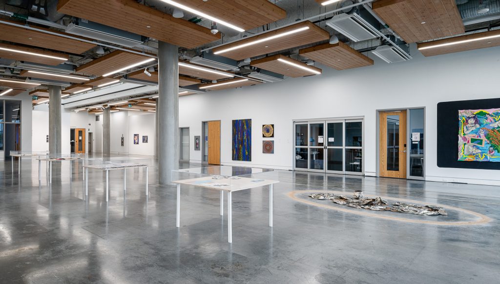 Interior view of Emily Carr University; in the foreground are several display tables topped with glass; in the background an installation of ceramic pieces lays on the floor encircled by light brown powder; in the background on the far wall hang colourful paintings out of focus. 
