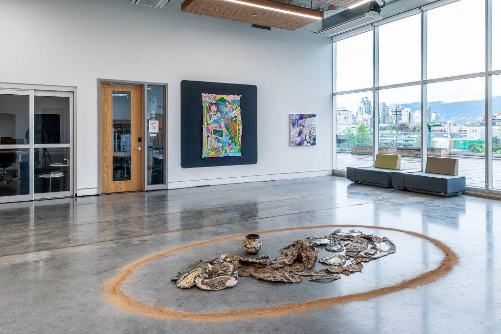 Inside view of Emily Carr University; in the foreground, an installation of flat textured brown ceramic pieces and one matching pot lays on the floor encircled by light brown powder. In the background are two colourful paintings; the one on the left is mounted on a larger black backdrop and shows girls amid plants; the one on the right is smaller and has many abstract shades of purple. 