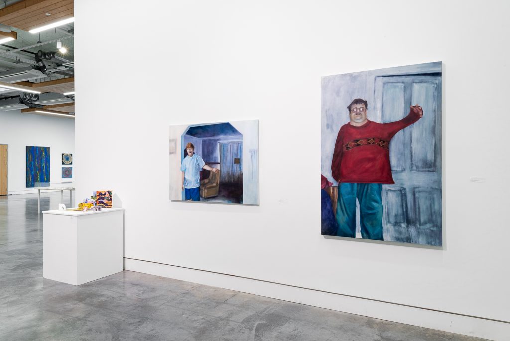 On the left mounted on a plinth are several colourful small pop-up cards and books. In the centre is a painting of a person with glasses and red hair wearing blue shorts and t-shirt, standing in a living room with their left arm raised. On the right is a painting of a person with glasses and dark hair wearing a red shirt and blue paints in front of a doorway with their left arm raised. 