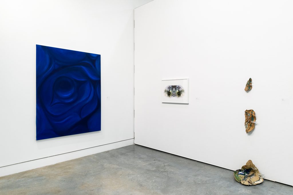 On the left wall, a large dark blue and black painting of an eye in Northwest Coast formline style; on the adjacent wall is a green and brown print with a white frame. Beside it are three irregular ceramic pieces that are arranged vertically so two are hung on the wall and one is on the floor. 