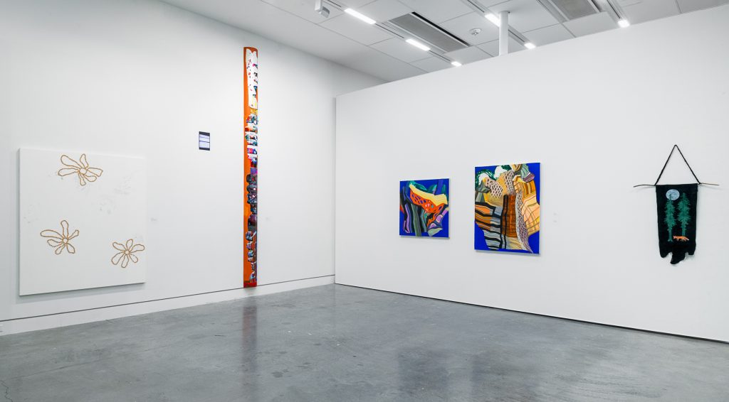On the left exhibition wall is a mostly-blank white canvas with outlines of brown flowers; beside it is a tall orange plank with colourful designs printed onto it. On the adjacent wall are two paintings with warped brown and yellow buildings on a cobalt background; beside is a black piece of hide hanging on the wall with an orange fox, white moon and green tree design. 