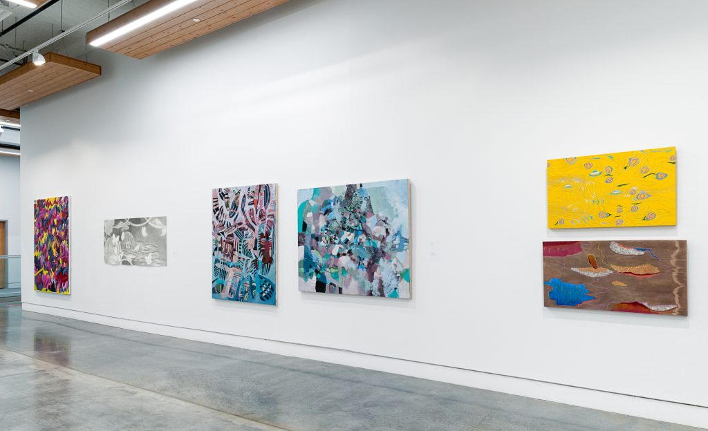 Six works on paper hang on an exhibition wall. The first is a busy abstract pink, black and yellow painting; the second is a charcoal drawing; the third and fourth are teal, periwinkle and lavender abstract paintings; the fifth is a yellow painting with green eye-like designs and the sixth is a brown painting with red, brown and blue patches. 