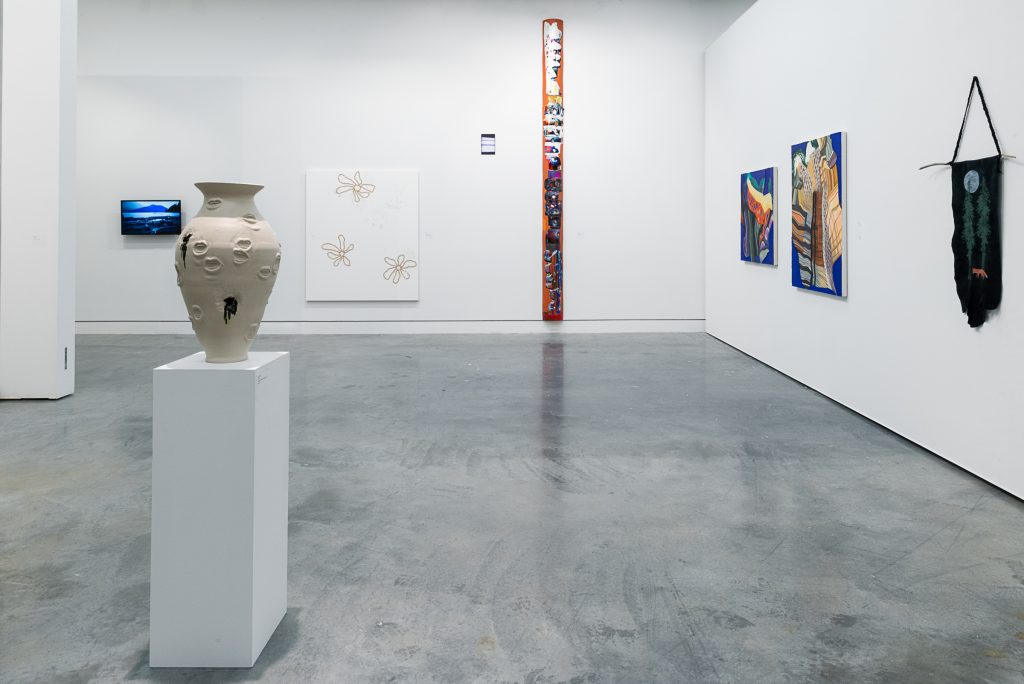 In an exhibition space, a cream-coloured ceramic vase with narrow neck and base sits on a plinth. Relief designs of mouths dot the vase’s surface. On the back wall is a TV screen showing a blue outdoor scene, a white canvas with brown outlines of flowers, and a tall orange plank printed with colourful designs. On the right wall are two paintings with abstract brown forms and cobalt blue backgrounds, and a black piece of hide hanging from a string with a moon, tree, and fox print. 