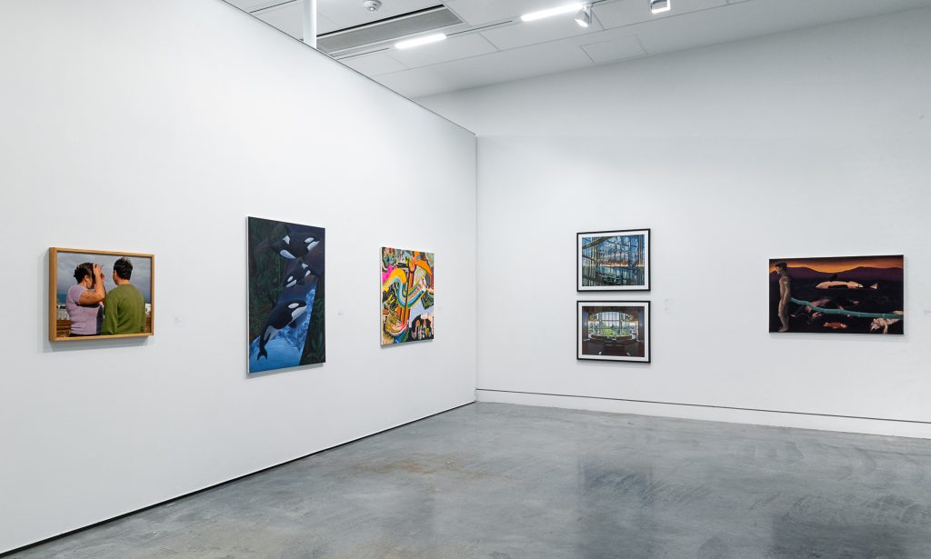Facing a corner, on the left exhibition wall is a wood-framed photograph of two people facing away from the camera, a dark blue and black painting of orca whales, and a colourful abstract painting with wide brushstrokes. On the adjacent right wall is a series of two framed photographs of a windowed room, and a brown painting of a brown-and-orange landscape with a standing human figure and animal carcasses on the ground. 