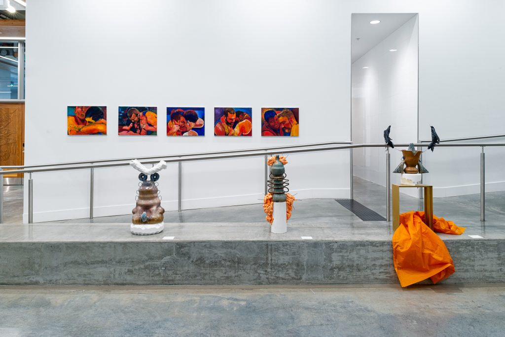 On a ledge beside an indoor ramp sits three industrial-looking ceramic sculptures with textured attachments. The rightmost sculpture sits on a wood stand containing an overflowing orange plastic tarp. On the far wall are a series of five close-up portraits of men wrestling painted in shades of orange and red, with dark blue backgrounds. 