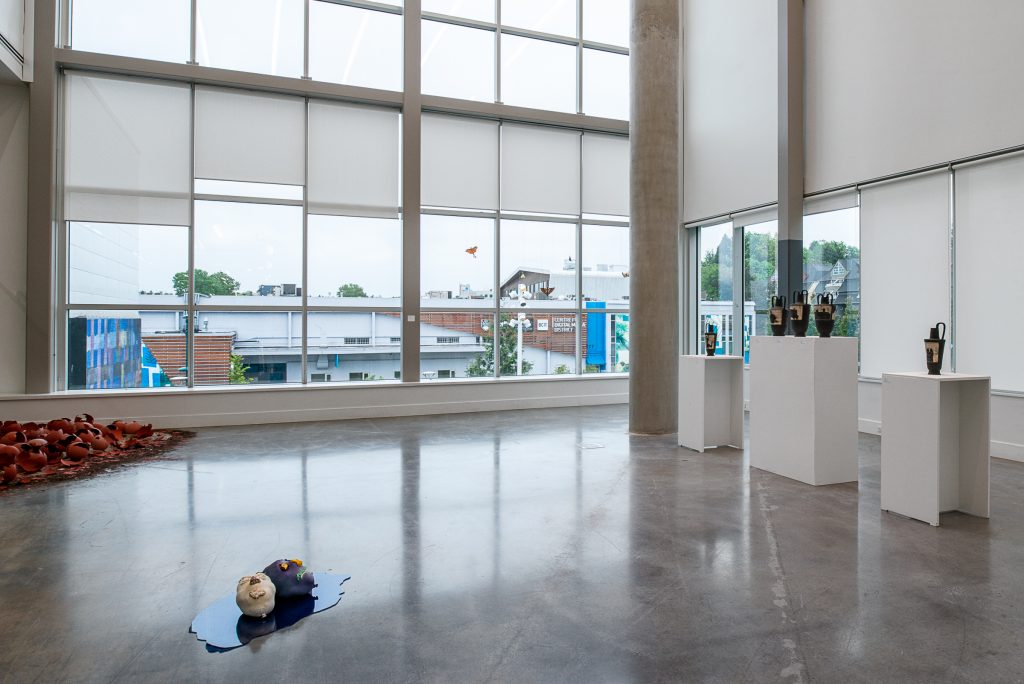 Facing a window, two small knobbly head sculptures sit on a reflective puddle-shaped surface on the ground. On the right wall are five black water jugs with brown etched patterns displayed on plinths, with the middle three jugs sharing one plinth. 