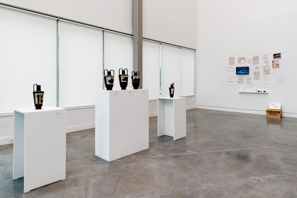 Five black water jugs with brown etched patterns are displayed on plinths, with the middle three jugs sharing one plinth. On the far wall is an installation of many pieces of brown and cream paper and some photographs. On the ground below is an upturned soap box with a placard atop it. 
