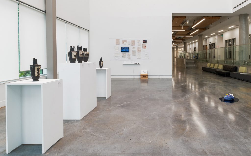 On the left, five black water jugs with brown etched patterns are displayed on plinths, with the middle three jugs sharing one plinth. On the far wall is an installation of many pieces of brown and cream paper and some photographs. On the ground is an upturned soap box with a placard atop it. To the right are two head-shaped sculptures sitting in a reflective puddle-shaped surface on the ground. 
