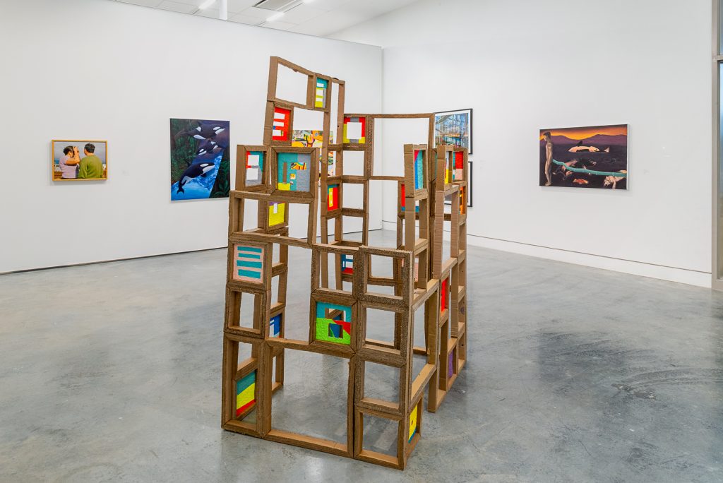 In an open exhibition space sits a square structure constructed from frames made of cardboard, with some of the frames filled with coloured inserts. On the far wall is a wood-framed photograph of two figures looking away from the camera, and a dark blue and black painting of orca whales. On the adjacent walls are two photographs blocked from view by the structure, and a brown and orange landscape painting with a standing human figure and slain animal carcasses on the ground.  