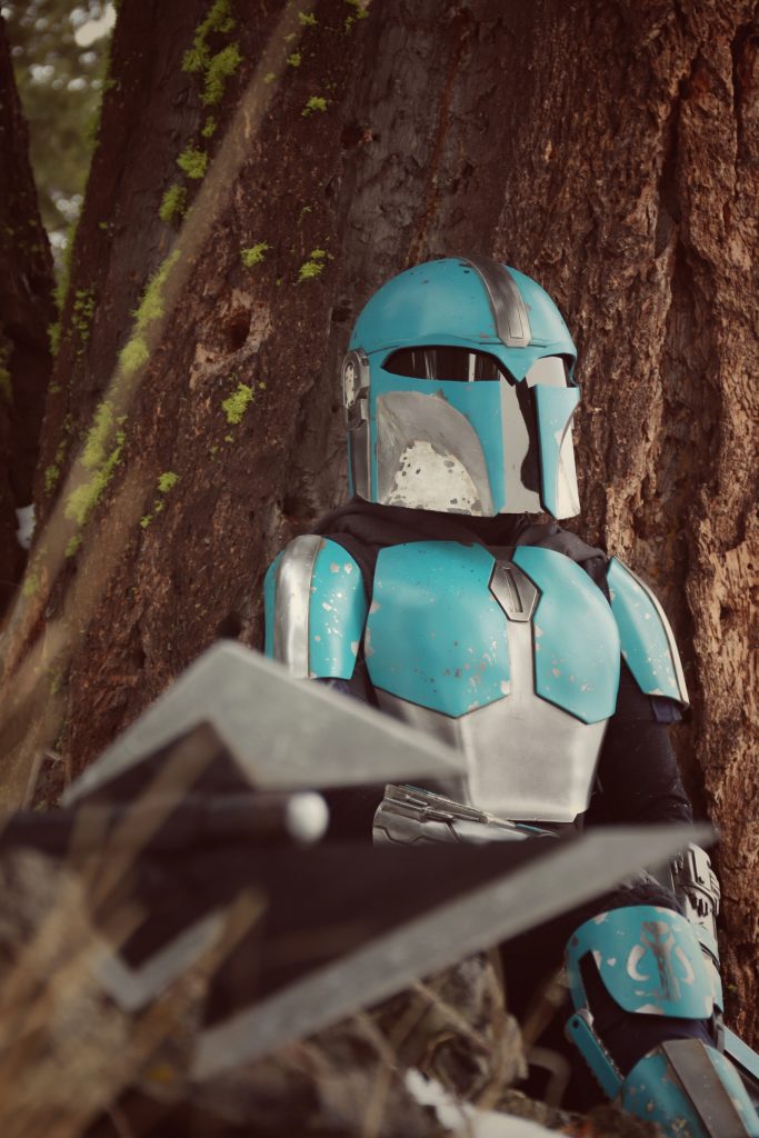 Person sitting against a big knobby tree. Wearing Mandalorian armour. Close up, with a spear in the foreground. 
