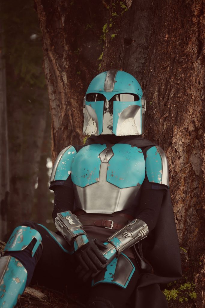 Person leaning back against a big knobby tree. Wearing Teal and Silver Mandalorian Armour, with arms crossed on their lap. 