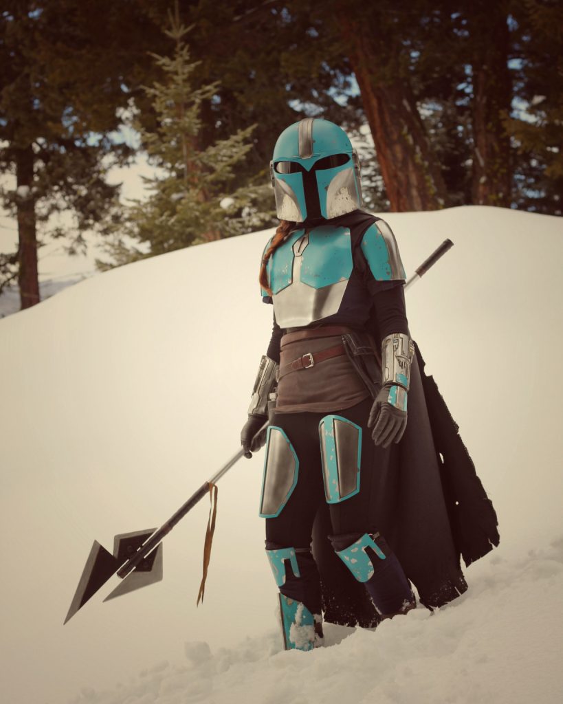 Person standing in shin deep snow on a mountain. Wearing Mandalorian armour, holding a spear downwards in their right hand. 