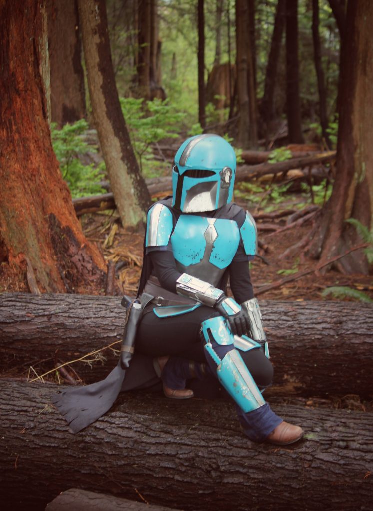 Person crouching on top of a big fallen log in the forest. Wearing armour looking over their shoulder. One hand placed on their knee. 