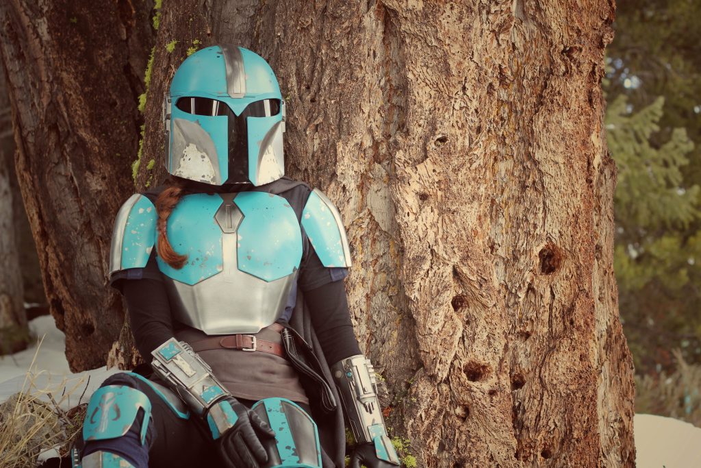 Person sitting against a big knobby tree. Wearing Teal and Silver Mandalorian Armour, with a helmet. Snow on ground. 