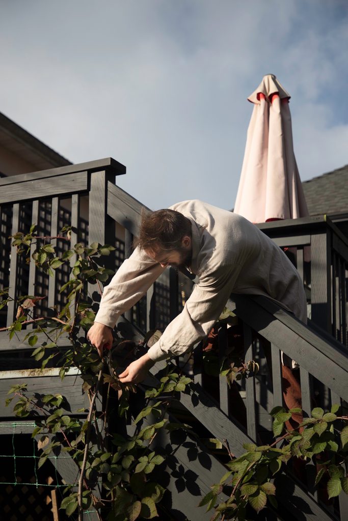 A person with short brown hair leans over a railing outside and trims a raspberry vine