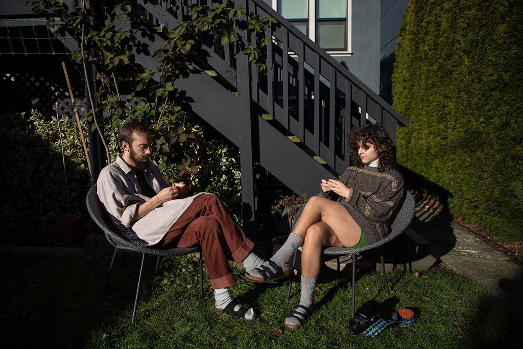 Two people, one with short brown hair and the other with shoulder-length brown hair each sit in a chair on the grass in front of a railing and peel grapefruit 