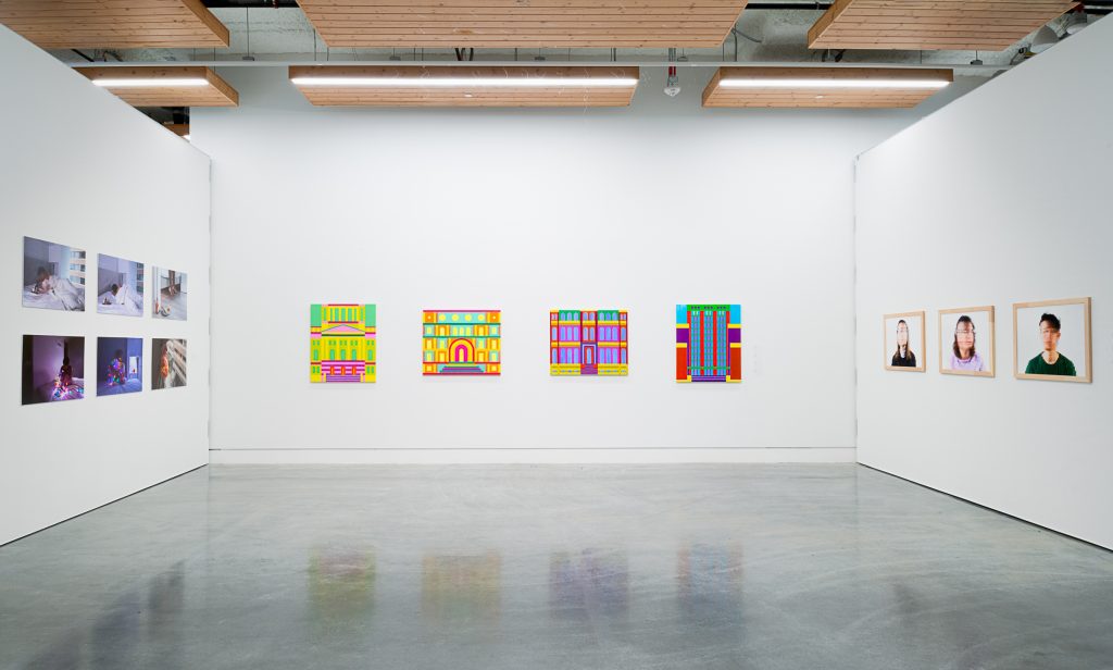 Four brightly coloured paintings of art deco building facades hang on a far wall. On the left wall are six photographs in purple tones. On the right wall are three wood-framed images of pixelated faces against white backgrounds. 
